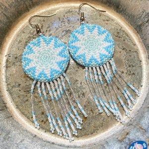 Peut inclure: Une paire de boucles d'oreilles en perles avec un motif en étoile bleu et blanc et des franges. Les boucles d'oreilles sont rondes et ont un crochet argenté.