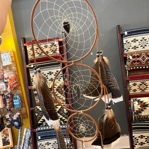 May include: A vertical arrangement of three circular dream catchers with brown wrapped hoops and cream-colored webbing. The structure features hanging brown and white feathers, dark beads, and a central black stone arrowhead in the top hoop.