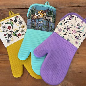 May include: Three oven mitts with silicone hands and fabric cuffs. The left mitt is mustard yellow with a hummingbird and floral print. The center mitt is turquoise with a forest, moose, and bear scene. The right mitt is purple with a bird pattern.