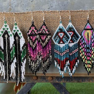 Pode incluir: Uma coleção de brincos de contas em vários designs geométricos e cores. Os brincos apresentam padrões intrincados com contas turquesa, vermelhas, pretas, brancas, rosa e verdes. Cada par tem uma forma e franjas únicas.