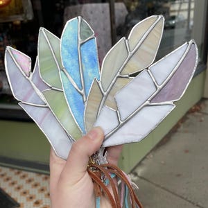 May include: A collection of five stained glass feathers in shades of blue, green, white, and lavender, joined by silver metal solder lines. The feathers are attached to a cluster of thin, dangling suede leather cords in brown, blue, and tan colors.