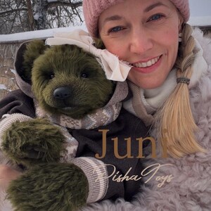 May include: A handmade olive green teddy bear wearing a brown hooded jacket, a patterned scarf, and a white bow. The bear is being held by a person wearing a pink beanie. The text "Juni Disha Toys" is visible.