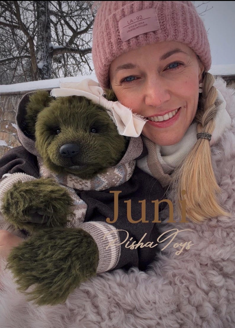 May include: An olive green teddy bear wearing a brown hooded jacket, a patterned scarf, and a white bow. The bear is held by a person wearing a pink beanie. The text "Juni Disha Toys" is visible.