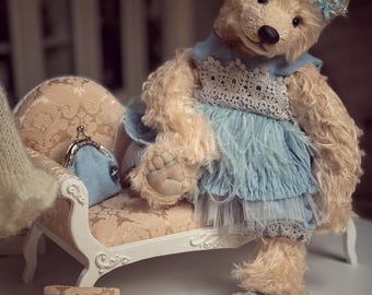 May include: A teddy bear dressed in a blue dress with lace detail and a small crown, seated on a miniature sofa. A matching purse and light blue slippers are also present. The rug is brown.