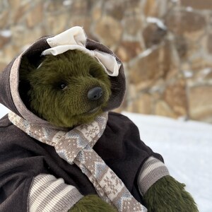 May include: A handmade olive green teddy bear wearing a brown coat with a hood and a cream-coloured bow. The bear has a patterned scarf and matching mittens. The background is a stone wall and snow.