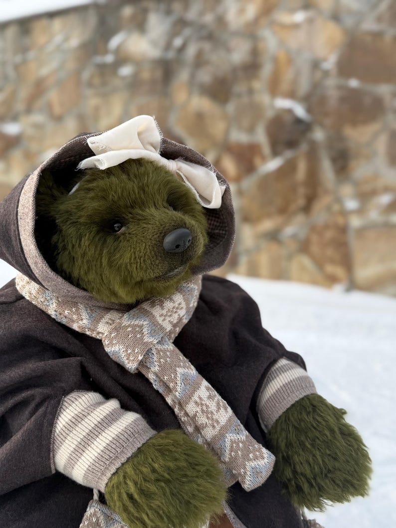 May include: A handmade olive green teddy bear wearing a brown coat with a hood and a cream-coloured bow. The bear has a patterned scarf and matching mittens. The background is a stone wall and snow.