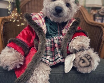 May include: A teddy bear dressed in a red coat with faux fur trim and a plaid dress, wearing a festive antler headband. The bear is seated on a green cushion. The bear's fur is light brown, and the coat has red and brown accents.