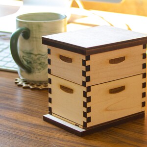 Puede incluir: Una caja de madera de dos niveles con una tapa marr&oacute;n oscuro y un borde negro. La caja est&aacute; hecha de madera clara y tiene un dise&ntilde;o sencillo.
