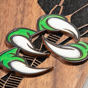 May include: Close-up of three green, white, and brown decorative pins. Each pin features a stylized design with green and white sections, outlined in brown. The pins are placed on a wooden surface with a dark pattern.