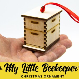 May include: A wooden beehive ornament with a red ribbon for hanging. The ornament is a miniature version of a traditional beehive with three stacked boxes. The ornament is labeled "My Little Beekeeper" and "Christmas Ornament".