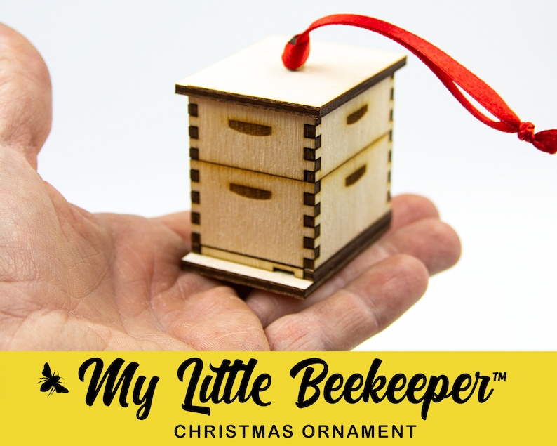 May include: A wooden beehive ornament with a red ribbon for hanging. The ornament is a miniature version of a traditional beehive with three stacked boxes. The ornament is labeled "My Little Beekeeper" and "Christmas Ornament".