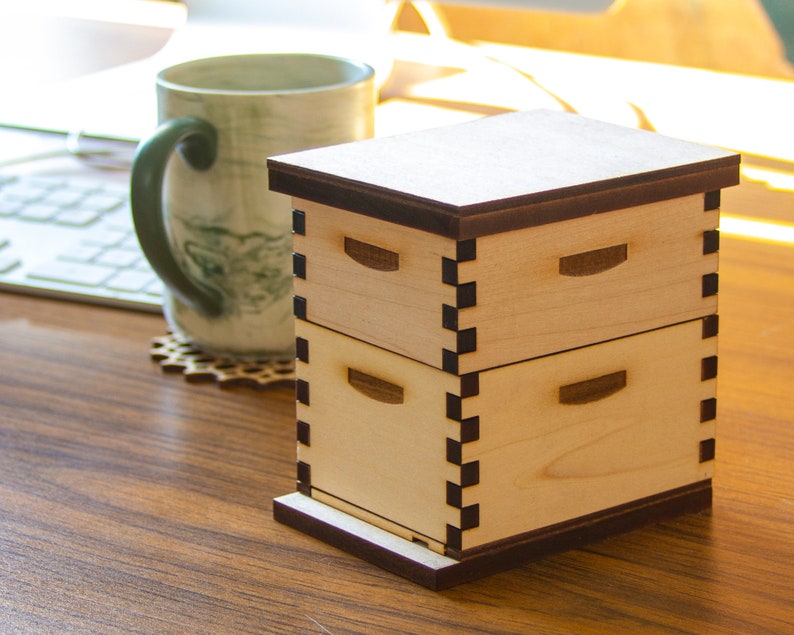 Puede incluir: Una caja de madera de dos niveles con una tapa marr&oacute;n oscuro y un borde negro. La caja est&aacute; hecha de madera clara y tiene un dise&ntilde;o sencillo.