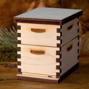 Puede incluir: Una peque&ntilde;a caja de madera con dos cajones, cada uno con una peque&ntilde;a manija recortada. La caja est&aacute; hecha de madera clara con ribetes marrones oscuros. La caja est&aacute; sentada sobre una superficie de madera.