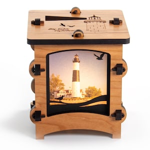Könnte beinhalten: Eine Holzkiste mit einer Leuchtturmszene auf der Vorderseite. Die Kiste ist hellbraun mit schwarzen Akzenten und hat einen Schlitz oben. Die Vorderseite zeigt ein Bild eines schwarz-weißen Leuchtturms mit Vögeln und dem Text "Big Sable Lighthouse".