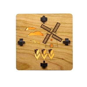 Puede incluir: Un adorno de pared de madera con un dise&ntilde;o de molino de viento, nubes y tulipanes cortado con l&aacute;ser. El molino de viento es marr&oacute;n y las nubes son naranjas. Los tulipanes son amarillos y verdes. El fondo es de madera clara.