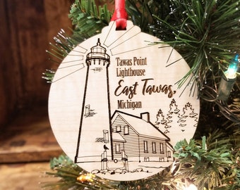 May include: A round wooden Christmas ornament with a laser-etched design of the Tawas Point Lighthouse in East Tawas, Michigan. The ornament has a red ribbon for hanging and is displayed on a Christmas tree.