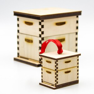 May include: A small wooden beehive ornament with a red ribbon for hanging. The ornament is made of light wood and has a natural finish. It is a miniature version of a traditional beehive.