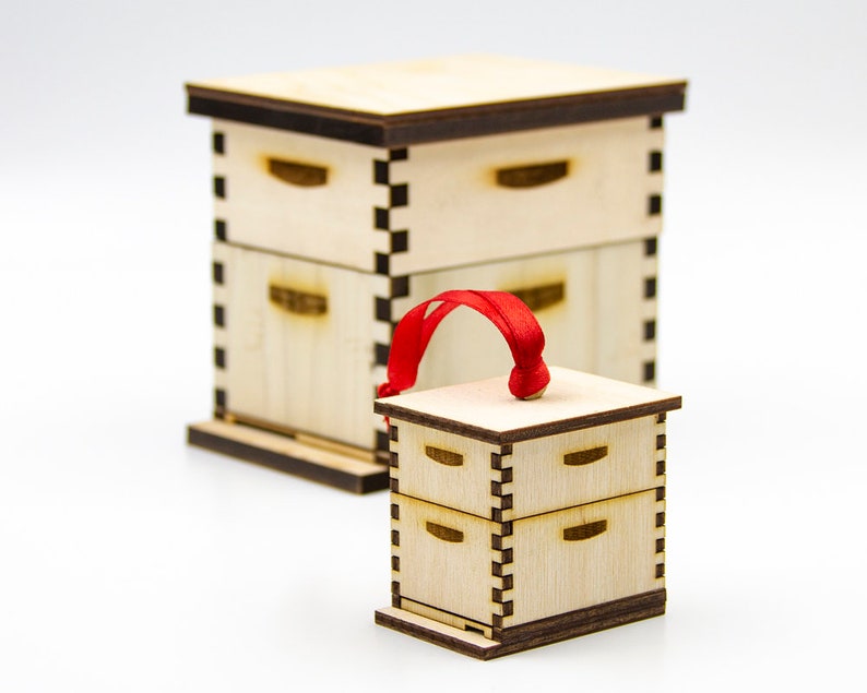 May include: A small wooden beehive ornament with a red ribbon for hanging. The ornament is made of light wood and has a natural finish. It is a miniature version of a traditional beehive.