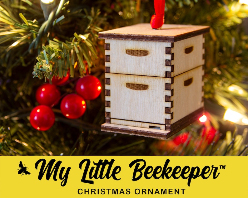 May include: A wooden Christmas ornament shaped like a beehive with two drawers. The ornament is hanging on a Christmas tree with red berries and lights.