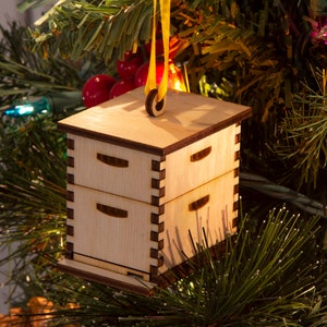 May include: A wooden Christmas ornament shaped like a beehive with three drawers. The ornament is hanging from a yellow ribbon on a Christmas tree.