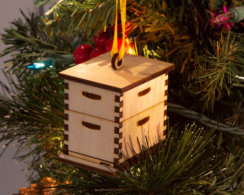 May include: A wooden Christmas ornament shaped like a beehive with three drawers. The ornament is hanging from a yellow ribbon on a Christmas tree.