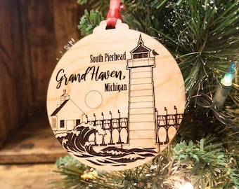 May include: A wooden Christmas ornament featuring a black and white illustration of the South Pierhead Lighthouse in Grand Haven, Michigan. The ornament is round and has a red ribbon attached to the top.