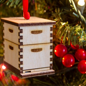 May include: A wooden beehive ornament with a red ribbon hanging from a Christmas tree. The ornament is made of light wood and has a brown base.