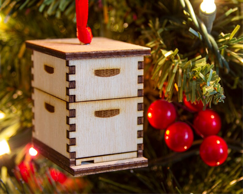 May include: A wooden beehive ornament with a red ribbon hanging from a Christmas tree. The ornament is made of light wood and has a brown base.