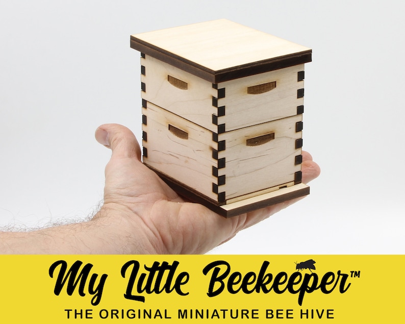 Puede incluir: Una colmena de abejas en miniatura de madera con dos cajas apiladas, una base marr&oacute;n y un dise&ntilde;o a cuadros en blanco y negro. La colmena se sostiene en la mano de una persona. El texto "My Little Beekeeper&trade; THE ORIGINAL MINIATURE BEE HIVE" se muestra debajo de la colmena.