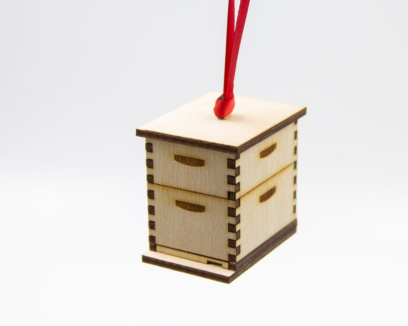 May include: A wooden beehive ornament with a red ribbon. The beehive is made of light wood and has two drawers. The ornament is perfect for hanging on a Christmas tree or other holiday decor.