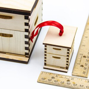 May include: A small wooden beehive ornament with a red ribbon for hanging. The ornament is about 2 inches tall and is made of light wood with dark brown trim. The ornament has two drawers and a flat top.