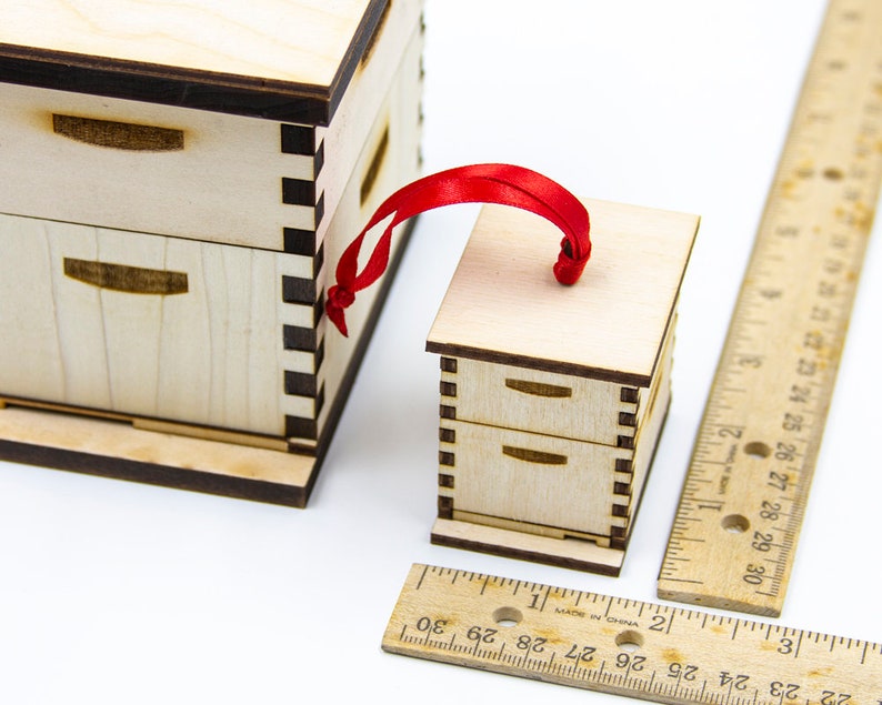 May include: A small wooden beehive ornament with a red ribbon for hanging. The ornament is about 2 inches tall and is made of light wood with dark brown trim. The ornament has two drawers and a flat top.