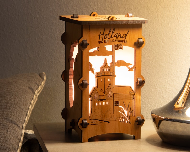 Puede incluir: Una l&aacute;mpara de madera con un dise&ntilde;o recortado del faro Big Red en Holland, Michigan. La l&aacute;mpara est&aacute; encendida y la luz brilla a trav&eacute;s del dise&ntilde;o recortado. La l&aacute;mpara est&aacute; sobre una mesa con una almohada y un jarr&oacute;n plateado.