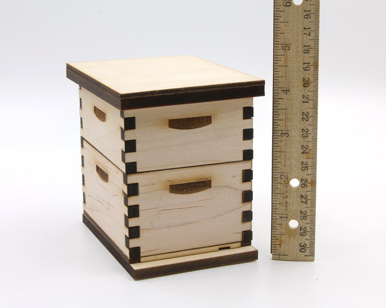 Puede incluir: Una colmena de madera en miniatura con dos cajones, que mide aproximadamente 15 cm de alto, se muestra junto a una regla. La colmena est&aacute; hecha de madera clara con molduras de color marr&oacute;n oscuro y detalles negros.