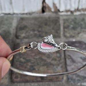 May include: Silver-toned bangle bracelet featuring a decorative charm. The charm has a pink and white enamel design with a silver mountain and is encrusted with small, clear crystals. The bracelet is held by a hand.