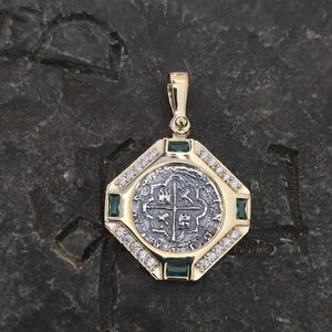 May include: A gold pendant with a silver coin at the center, featuring a cross design. The pendant is surrounded by small diamonds and emeralds, with a gold bail at the top. The pendant is set against a dark, textured background.