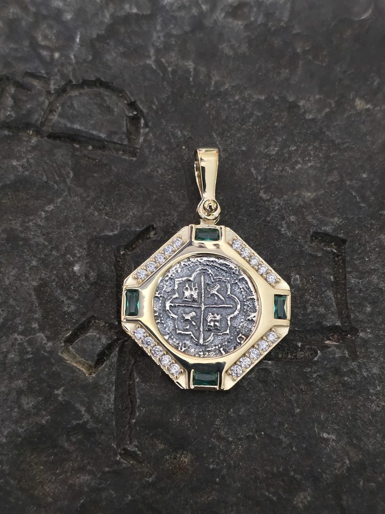 May include: A gold pendant with a silver coin at the center, featuring a cross design. The pendant is surrounded by small diamonds and emeralds, with a gold bail at the top. The pendant is set against a dark, textured background.