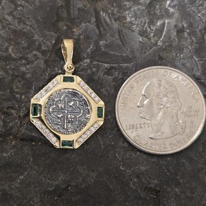May include: A gold pendant with a silver coin center, surrounded by diamonds and emeralds. The pendant is octagonal and has a bail for a chain. A U.S. quarter is next to the pendant for size comparison.