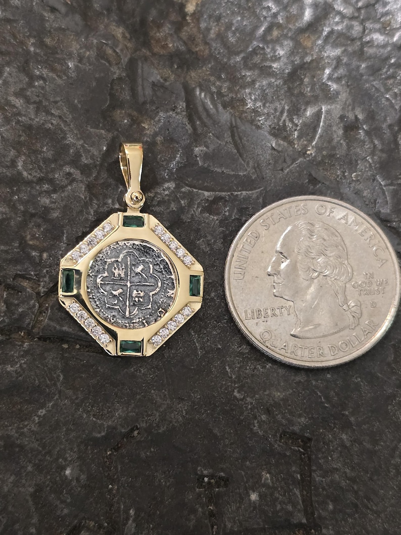 May include: A gold pendant with a silver coin center, surrounded by diamonds and emeralds. The pendant is octagonal and has a bail for a chain. A U.S. quarter is next to the pendant for size comparison.
