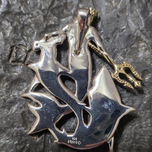 May include: A silver-toned pendant featuring a stylized depiction of a merman holding a trident. The merman is adorned with a crown and is surrounded by a stylized aquatic design. The pendant is marked with "925" and "Spirit".