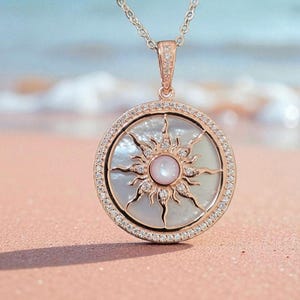 May include: Rose gold pendant necklace featuring a sunburst design. The pendant has a mother-of-pearl background with a sun design and a central pink stone. The pendant is 1.5 inches in diameter and is encrusted with small clear stones.