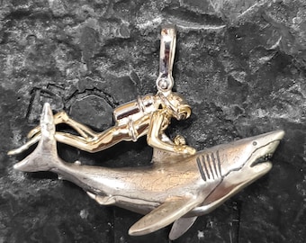 May include: A silver shark pendant with a gold diver riding on top. The diver is wearing a scuba tank and has a diving mask on.