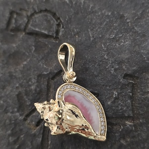 May include: A gold-colored pendant in the shape of a seashell. The pendant features a pink and white shell design, accented with small, clear gemstones. The seashell pendant is suspended from a gold bail.