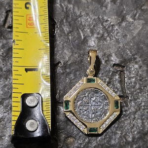 May include: Gold pendant with a silver coin center, surrounded by a gold octagonal frame. The frame is embellished with small diamonds and emeralds. A yellow measuring tape is next to the pendant.