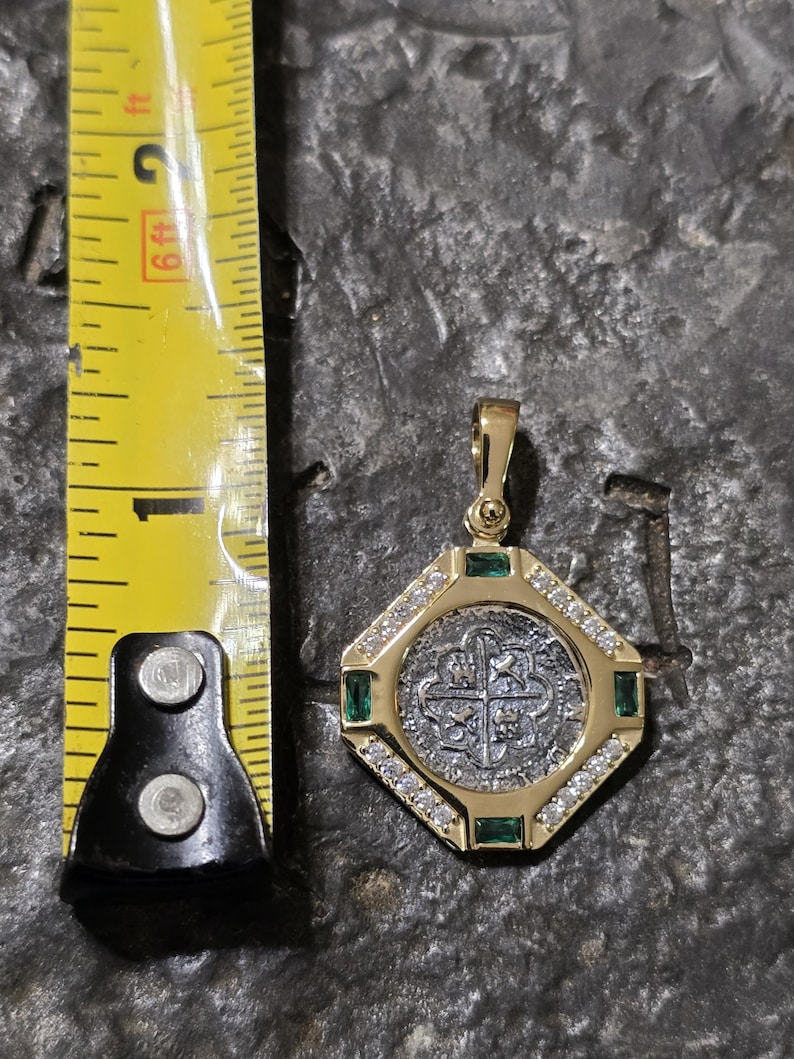 May include: Gold pendant with a silver coin center, surrounded by a gold octagonal frame. The frame is embellished with small diamonds and emeralds. A yellow measuring tape is next to the pendant.
