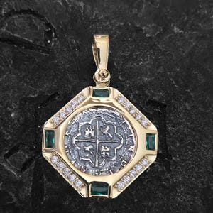 May include: A gold pendant with a silver coin center, emerald accents, and small clear stones. The pendant has a rectangular shape with rounded corners and a gold bail for a chain. The coin features a cross design.