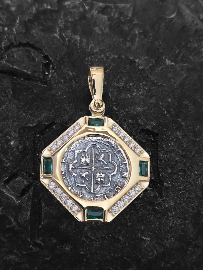 May include: A gold pendant with a silver coin center, emerald accents, and small clear stones. The pendant has a rectangular shape with rounded corners and a gold bail for a chain. The coin features a cross design.