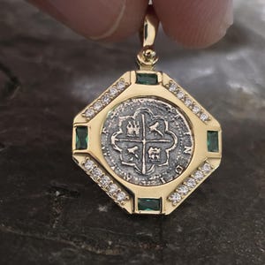 May include: A gold pendant featuring a silver coin with a cross design at the center, surrounded by a gold frame. The frame is embellished with small diamonds and green emeralds. The pendant is held by a person's fingers.