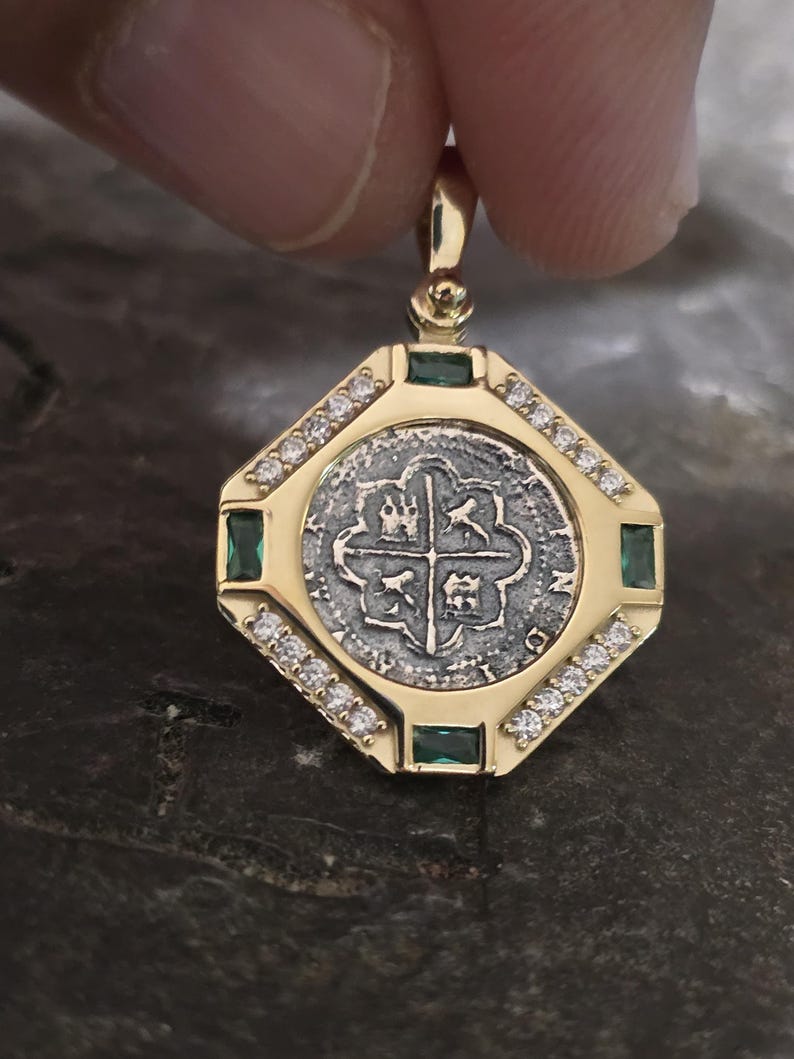 May include: A gold pendant featuring a silver coin with a cross design at the center, surrounded by a gold frame. The frame is embellished with small diamonds and green emeralds. The pendant is held by a person's fingers.