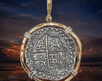 Op de afbeelding: Een gouden en zilveren hanger met een replica van een Spaanse acht reales munt. De munt heeft een kruis met een kasteel aan elke kant en een lelie boven en onder. De hanger hangt aan een gouden ketting.